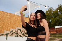 Elegantes amigos alegres en sombrero negro abrazando y tomando selfie en el teléfono móvil en el día brillante en la arena decorada en el festival - foto de stock
