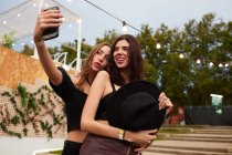 Stylish cheerful friends in black hat embracing and taking selfie on mobile phone in bright day at decorated arena on festival — Stock Photo