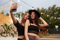 Eleganti amici allegri in cappello nero che abbracciano e prendono selfie su telefono cellulare in giorno lucente a arena decorata su festival — Foto stock