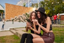 Pleasant cheerful friends in black hat having fun watching photo on mobile phone sitting on green lawn near decorated stage in bright day at festival — Stock Photo