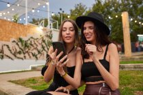 Friends in black hat having fun watching photo on mobile phone sitting on green lawn near decorated stage in bright day at festival — Stock Photo