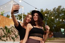 Elegantes amigos alegres en sombrero negro abrazando y tomando selfie en el teléfono móvil en el día brillante en la arena decorada en el festival - foto de stock