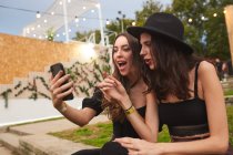 Друзья в черной шляпе весело смотрят на фото на мобильный телефон, сидя на зеленой ленточке возле украшенной сцены в яркий день на фестивале — стоковое фото