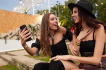 Friends in black hat having fun watching photo on mobile phone sitting on green lawn near decorated stage in bright day at festival — Stock Photo