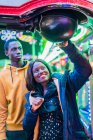 Feliz mujer negra sonriendo y golpeando la pelota mientras juega juego de arcade con el novio por la noche en la feria - foto de stock