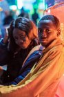 Side view of African American man looking at camera while sitting near girlfriend during date at night on funfair — Stock Photo