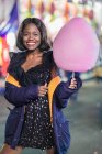 Ottimista femmina nera con filo interdentale caramelle sorridente e guardando la fotocamera mentre si diverte sul luna park di notte — Foto stock