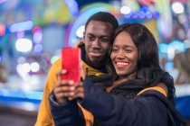 Joyeux homme et femme afro-américain souriant et prenant selfie pendant la date sur funfair la nuit — Photo de stock
