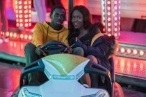 African American man and woman smiling and riding bumper car during date at night on fairground — Stock Photo