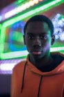 Young black man in orange hoodie looking at camera while standing against colorful lights on fairground — Stock Photo