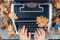 Hands of woman typing on vintage typewriter with tablet in autumn leaves on stone table in oak forest — Stock Photo