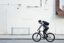 Side view of contemporary male cyclist in black helmet and eyeglasses surfing on mobile phone on white wall background — Stock Photo