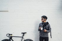 Сучасний чоловічий велосипедист у чорному шоломі та окулярах серфінгує на мобільному телефоні та п'є каву, щоб йти з білою стіною на фоні — стокове фото