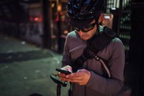 Contemporary male cyclist in black helmet and eyeglasses surfing on mobile phone on the street by night time — Stock Photo