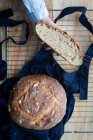 Vue du dessus de la main d'une femme anonyme tenant une tranche de pain au levain maison et une miche de pain sur un tablier sur un treillis de four . — Photo de stock