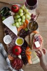 Von oben gesichtslose beschnittene Hände, die Snacks aus einem hölzernen Tablett mit Fleischstücken, Gemüsefrüchten und einem Glas Rotwein essen — Stockfoto