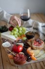 Зверху безликі обрізані руки їдять закуски з дерев'яного підносу з нарізаними м'ясними овочами фруктами та келихом червоного вина — стокове фото