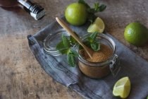 Açúcar mascavo suspenso em um pote perto de limas frescas e folhas de hortelã-pimenta colocadas em guardanapo em uma mesa de madeira — Fotografia de Stock