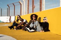 Focalizzato giovani donne diverse in abiti alla moda e occhiali da sole guardando la fotocamera mentre seduto durante la riunione amichevole sullo stadio — Foto stock