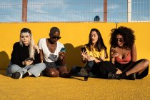 Mujeres jóvenes y diversas descuidadas en mensajes informales de ropa en el teléfono celular mientras están sentadas en el suelo de asfalto en el estadio - foto de stock