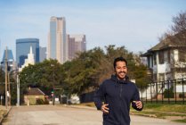 Happy Hispanic male athlete in sportswear with headphones running along empty road at downtown in Dallas — Stock Photo