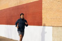 Vue latérale de fit homme sportif hispanique en tenue active écouter de la musique tout en faisant du jogging le long de la rue de la ville à Dallas — Photo de stock