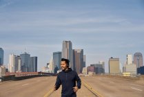 Corredor masculino hispano con capucha casual usando auriculares mientras corre con el cielo azul sobre el centro de Dallas, Texas - foto de stock