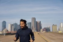 Joggeur hispanique masculin en sweat à capuche décontracté en utilisant un casque tout en courant avec ciel bleu au-dessus du centre-ville de Dallas, Texas — Photo de stock
