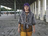 Модна стильна жінка з фіолетовою зачіскою в жовтих штанях дивиться в камеру на вулиці — стокове фото