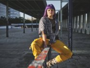 Stilvolle Frau mit leuchtend lila Frisur in gelben Hosen sitzt auf Metallzaun in der City Station und blickt in die Kamera — Stockfoto
