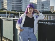 Fashion stylish woman with purple hairstyle touching face and leaning on metal fence in city center — Stock Photo