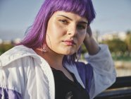 Fashion woman with purple hairstyle touching face and leaning on metal fence in city center and confidently looking in camera — Stock Photo