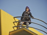 Низкий угол уверенной женщины с фиолетовой прической в блестящей черной куртке и солнцезащитных очках, смотрящей в сторону города — стоковое фото