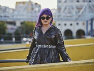 Femme confiante à la mode avec coiffure violette en veste noire brillante et lunettes de soleil regardant à la caméra sur le point de vue de la ville — Photo de stock