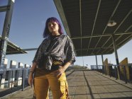 Stylish woman with purple hairstyle in yellow pants standing in street and looking in camera — Stock Photo