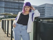 Fashion woman with purple hairstyle touching face and leaning on metal fence in city center and confidently looking in camera — Stock Photo