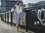 Стильна жінка з фіолетовою зачіскою в модному одязі позує в міському мосту в місті — стокове фото