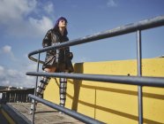 Уверенная женщина с фиолетовой прической в блестящей черной куртке, смотрящая на город с металлическим забором — стоковое фото
