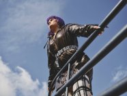 Низкий угол прически женщины с фиолетовой прической в блестящей черной куртке, смотрящей в сторону города на голубое небо — стоковое фото