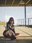 Giovane donna in occhiali da sole con acconciatura viola in giacca nera lucida comodamente seduta su asfalto con gambe incrociate in città — Foto stock