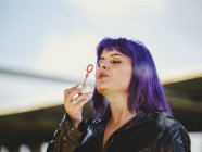 Porträt einer modischen Frau mit lila Haaren, die mit gepflegter Hand Blasen weht — Stockfoto
