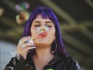 Ritratto di donna alla moda con capelli viola che soffia bolle con mano curata — Foto stock