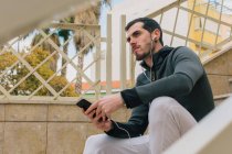 From below of pensive young man in tracksuit with earphones messaging on mobile phone taking break sitting on stairs looking away — Stock Photo