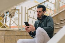 From below of pensive young man in tracksuit with earphones messaging on mobile phone taking break sitting on stairs — Stock Photo