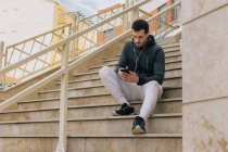 Снизу задумчивый молодой человек в спортивном костюме с наушниками, обменивающимися сообщениями на мобильном телефоне, отдыхающий на лестнице — стоковое фото