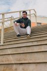 Знизу стомлений молодий чоловік у костюмі з навушниками на мобільному телефоні робить перерву, сидячи на сходах — стокове фото