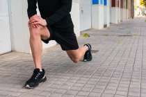 Безликий бегун в спортивной одежде разминает ноги, опираясь на колено, готовясь к пробежке на городской улице. — стоковое фото