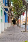 Vue arrière du gars actif en vêtements de sport avec écouteurs avec téléphone dans la main jambes d'entraînement jogging dans la rue urbaine — Photo de stock
