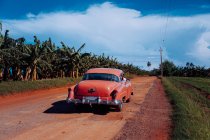 Sandy dirt red road with old vintage car with green plants on sides and grey cloudy sky on background in Cuba — Stock Photo