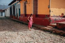 Mujer de vacaciones en vestido rosa claro y mochila caminando por un camino empedrado vacío entre viejos edificios coloridos en Cuba - foto de stock